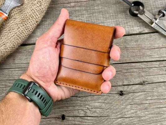 Hand holding a minimalist genuine leather wallet on a rustic wooden table with tools in the background.