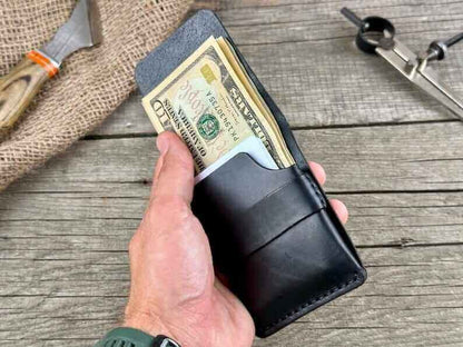 A hand holding a minimalist genuine leather wallet with cash visible inside on a rustic wooden surface.